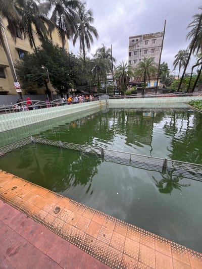 The image depicts an outdoor scene in Sylhet, Bangladesh, featuring a large, rectangular pool of murky green water. The pool is surrounded by a tiled walkway. In the background, several buildings of varying heights are visible, some appearing old and weathered, others more modern. Coconut palm trees are scattered throughout the scene, indicating a tropical or subtropical climate. A few people are present near the pool, some standing and observing, others seated. There's a metal fence submerged in the water, dividing a section of the pool. The sky is overcast, suggesting cloudy weather. The overall impression is that of a public space, possibly a park or a communal area, with the pool being a central feature.