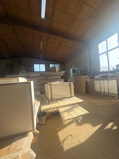 The image depicts the interior of a workshop, likely a furniture manufacturing facility, located in the city of Belene, Bulgaria. The space is filled with stacks of wood panels and pre-fabricated furniture components, many of which are covered in protective plastic wrap. Natural light streams in from large windows on the right side of the frame, casting strong shadows across the concrete floor. Several fluorescent lights are visible on the wooden ceiling, providing artificial illumination. The overall impression is one of an active workspace with materials and unfinished products organized for assembly or storage. There are no people visible in the image.