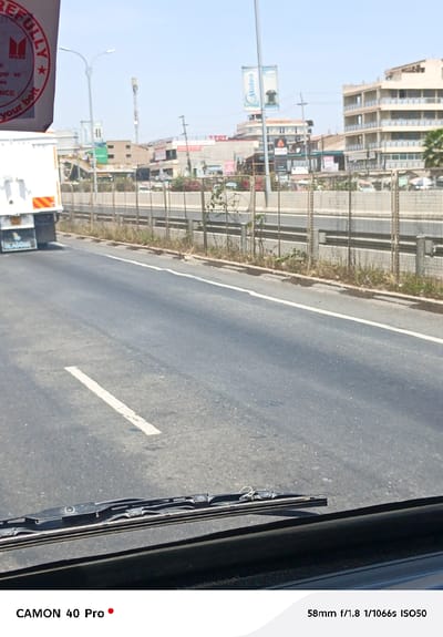 The image presents a bright, sunlit view from the interior of a vehicle, looking out through the windshield onto a multi-lane road. The windshield wipers are visible at the bottom edge of the frame, indicating the perspective is from the driver or front passenger seat.
To the left, a large white truck or lorry is partially visible, moving in the same direction or stopped alongside. On its rear mud flap, the word "BLABONG" is discernible. A red circular sticker on the back of the truck features the "ISUZU" logo and partially visible text, including "REFULLY" (likely part of "CAREFULLY") and "your belt", suggesting a safety message.
The asphalt road surface shows wear and tear, with clear white lane markings โ a solid white line defining the shoulder on the right and a faded dashed white line in the center of the visible lanes. A low metal fence or barrier separates the immediate roadway from a parallel, lower-lying section of road or undeveloped land, beyond which numerous buildings line the horizon.
The background reveals a bustling urban or semi-urban environment under a clear, bright blue sky, indicative of a sunny day, likely midday or early afternoon given the high sun and minimal shadows. Several commercial buildings are visible, displaying various signs. Notably, a prominent sign for "Midea" is seen on one building, and the letters "TTOT" are visible on another building's signage. A tall, slender communication mast or antenna stands out against the sky in the distance. Streetlights with double luminaires are also scattered throughout the scene.
The overall impression is that of a typical day on a busy road in a developed area. The image's metadata, visible at the bottom, indicates it was captured with a "CAMON 40 Pro" phone, using settings of "58mm f/1.8 1/1066s ISO50".