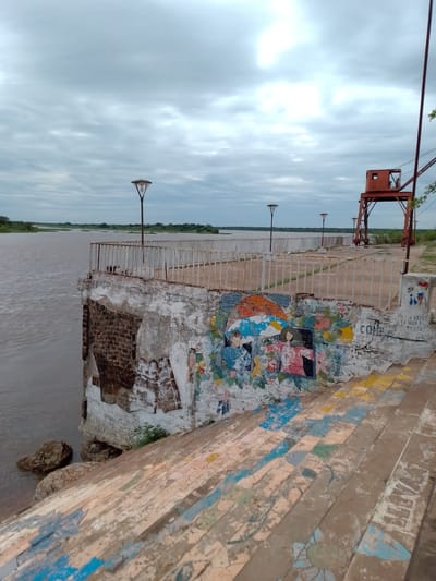The image depicts a tranquil riverside scene in Concepción, Paraguay, under a heavily overcast sky. A wide, muddy-brown river occupies the left side, with distant green vegetation lining its far bank. On the right, a concrete embankment or pier extends into the distance, featuring a white metal railing and several classic lampposts with distinct tops.

In the upper right background, a prominent, rusty red industrial crane structure stands, suggesting a past or present port function. The wall beneath the pier, facing the viewer, exhibits crumbling brickwork and faded plaster. This wall is adorned with a vibrant, multi-colored mural in a folk-art style, depicting figures, umbrellas, flowers, and abstract patterns. Below this, a wide set of concrete steps descends towards the river, with each tread painted with colorful, geometric, and floral designs that mimic a mosaic. Large rocks are visible at the base of the embankment, partially submerged in the river.

No people are visible in the scene. The cloudy weather creates a soft, diffused light, indicating a dull day. Visible text on the mural wall includes "CORR" and "SOST". On a small concrete block near the crane, faint Spanish text reads "QUE DUELE", "LA PARA DE", and "2018", accompanied by a small drawing of two figures. The overall impression is a blend of historical industrial elements, natural beauty, and local artistic expression in a public waterfront setting.