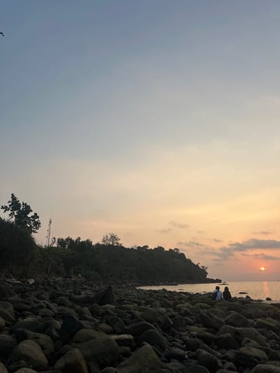 A coastal scene is depicted at sunset in the city of Preah Sihanouk, Cambodia. The foreground consists of a dense field of dark, irregularly shaped rocks forming a beach. In the middle-ground, two individuals are seated on these rocks, facing the ocean with their backs partially to the viewer; one wears a light-colored top, and the other wears a dark top. The calm ocean surface reflects the sky's colors. The sun, a distinct orange-red disc, is positioned just above the ocean horizon on the right side of the frame. A dark, densely forested hill or cliff line borders the coast in the background. A tall, slender communication tower is visible rising from the left side of this hillside. The sky exhibits a gradient from light blue overhead to warm orange and yellow hues at the horizon, with scattered clouds. A small, dark, bird-like silhouette is visible in the upper left quadrant of the sky. The overall lighting indicates twilight, casting foreground elements into silhouette.