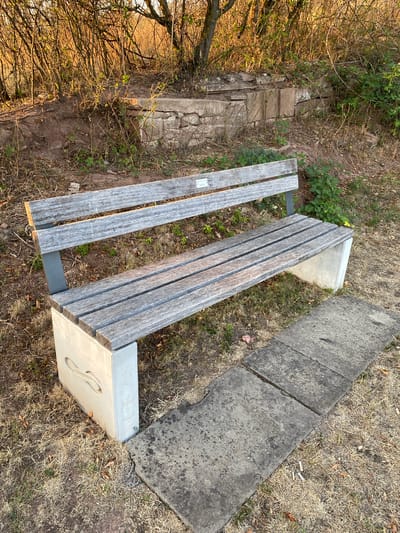 A weathered wooden park bench with concrete supports sits on dry grass in front of a low, rough-hewn stone wall overgrown with weeds and thorny bushes. The bench is constructed of parallel wooden slats for both the seat and the backrest. One of the concrete supports features a carved decorative motif resembling a double loop or infinity symbol. To the right of the bench, a section of concrete paving slabs extends into the frame. The scene appears to be outdoors, possibly in a park or along a trail, with dry vegetation suggesting a warm or dry climate. Sunlight casts long shadows, indicating it is either early morning or late afternoon. The image was taken in Waiblingen, Germany.