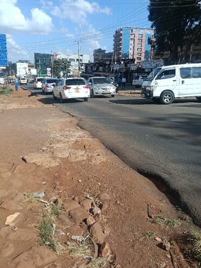 The image displays a daytime urban street scene in Ndenderu ward, Kenya, under a sky with scattered clouds. The foreground consists of an unpaved, reddish-brown dirt shoulder, severely eroded along its edge, creating a distinct drop-off to the asphalt road. This shoulder contains scattered rocks, rubble, and patches of sparse green weeds, with some visible debris.
The asphalt road, occupying the middle ground, carries traffic. A white Toyota Hiace-style van is positioned in the right foreground lane. Behind it, a silver sedan (license plate KDE 404H) is visible, followed by several white station wagons, including one with license plate KDG 192A and another with KDG 455D. These vehicles appear to be in active traffic flow or queued.
On the left side of the road, an individual in a blue shirt walks on the unpaved shoulder. On the right, a group of approximately three to four people stands near commercial buildings, possibly on a sidewalk. The background features a dense concentration of multi-story buildings, including a prominent structure with blue reflective glass panels. Overhead utility poles and numerous power lines crisscross the sky. Various commercial signs, such as "DIGITAL SOLUTIONS" and "HAVEN," are visible on the building facades.