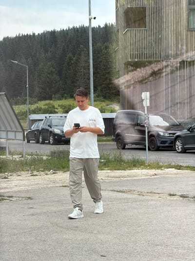 A man in a white t-shirt and tan cargo pants is walking on an asphalt path, looking at his phone. He is wearing white sneakers. Behind him, several cars are parked, including a black SUV, a dark gray van, and a dark gray sedan. In the background, a dense forest covers a hillside, and a modern building with a wooden facade stands to the right. A security camera is visible on a pole in the foreground. The location is Žunovnica, Bosnia and Herzegovina.