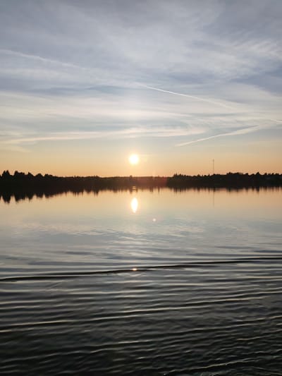 A serene body of water, likely a lake or river, reflects the warm hues of a sunset. The sky is a blend of soft blues and oranges, with wispy white clouds and contrails stretching across it. A bright, round sun is setting near the horizon, casting a golden glow on the water. The opposite bank is lined with a dense silhouette of trees, with a tall, thin communication tower visible on the right side. The foreground of the water exhibits ripples, indicating gentle movement, with a distinct wake pattern in the lower right portion of the image. No people or animals are visible in the scene. The overall impression is one of natural tranquility and the quiet beauty of a sunset over water. The image is not specific to the city of Bol'shaya Stepanovskaya, Russia, as there are no identifiable landmarks or geographical features that would confirm its location.