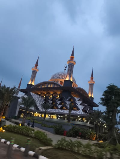 This image depicts a striking, modern mosque at dusk or early night, located in Cibinong, Indonesia. The sky is a deep, cloudy blue-grey, providing a dramatic backdrop for the illuminated structure.
The mosque features a prominent central dome adorned with intricate patterns and topped with a crescent moon finial. Multiple slender minarets, each with a pointed spire and illuminated band, rise from different points of the mosque. The building's main body showcases a complex, multi-tiered roof system composed of numerous interconnected geometric or triangular facets, also displaying elaborate patterns. Warm, golden light spills from within the mosque and illuminates its exterior details, highlighting its unique architectural style.
In the foreground and surrounding the mosque is lush green landscaping, including palm trees and various bushes. A paved pathway is visible in the lower left, where two figures, appearing to be individuals in traditional long garments, are walking away from the viewer. A black and white striped barrier or curb runs along the very bottom left edge of the frame, suggesting a roadside or designated pathway. The overall scene captures the mosque's grandeur and its contemporary design, beautifully accented by the evening illumination against the twilight sky. No visible text is clearly discernible in the image.