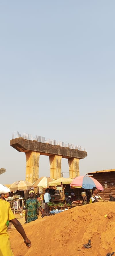 An unfinished concrete overpass looms over a bustling market scene. Sturdy pillars and a broad top with protruding rebar suggest future construction. Below, market stalls shaded by colorful umbrellas are filled with people and goods. A large mound of reddish-brown earth dominates the foreground, obscuring some of the activity. People are seen walking and interacting around the stalls, creating a vibrant, everyday scene juxtaposed against the incomplete infrastructure.