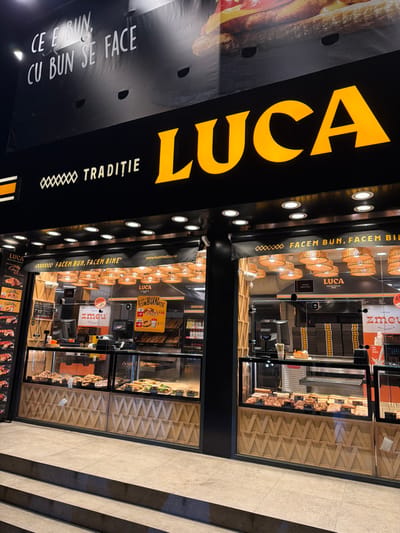 This image depicts the brightly illuminated storefront of a "LUCA TRADIȚIE" commercial establishment, presumably a bakery or pastry shop, located in Agigea, Romania, during nighttime hours. The prominent "LUCA" sign is rendered in large, yellow, three-dimensional letters on a black facade, with "TRADIȚIE" in white lettering to its left. Above the main sign, a large black banner features white text reading "CE E BUN, CU BUN SE FACE" and a partial image of a large sandwich or burger.

The storefront consists of multiple large glass display windows that reveal the store's interior. Inside, numerous woven, basket-like pendant lights illuminate the space. Below the windows, the lower portion of the storefront is adorned with a light-colored wooden paneling featuring a geometric, zigzag pattern.

Visible through the glass, multiple tiered glass display cases are filled with various baked goods, pastries, sandwiches, and other food items. Further inside, cash registers or service counters are discernible, alongside promotional signage, including one mentioning "ZMEU" and an offer for "1 COF BUNĂTĂȚI." Stacks of black packaging boxes are also visible. No individuals are present or clearly visible within the frame, either inside the store or on the exterior sidewalk. The immediate exterior ground features light-colored paved tiles, with two concrete steps leading up to the entrance level of the store.