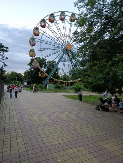 This image depicts a park scene in Kelermesskaya, Russia, featuring a large Ferris wheel as the central attraction. The Ferris wheel, with its green metal structure and numerous gondolas, dominates the upper portion of the frame. The gondolas are painted in red and yellow, and some appear to be occupied.
In the foreground and midground, a paved walkway made of rectangular bricks in shades of beige and brown runs diagonally from the lower left to the mid-right of the image. Several individuals are walking along the path. On the left side of the walkway, a group of young women is strolling, some looking towards the camera. Further down the path, an older man is pushing a stroller.
To the right of the walkway, a grassy area is visible, dotted with trees and bushes. A dark green tree with dense foliage partially obscures the Ferris wheel on the right side of the image. In the mid-right, a family is seated on a bench. A woman in a light blue top and white shorts is standing near them, and a black stroller is parked beside the bench. A young boy in a blue shirt stands in front of the bench.
In the background, more trees and a building, possibly a ticket booth or small kiosk, are present. The sky above is overcast with scattered clouds, suggesting it is either early morning or late afternoon. The overall atmosphere is that of a leisure park, with visitors enjoying the evening.