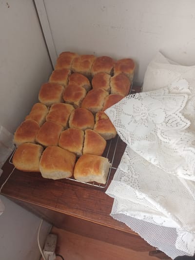 The image shows a metal cooling rack filled with freshly baked bread rolls. The rolls are golden brown and appear to be soft. They are arranged in rows on the rack. To the right of the cooling rack, a white lace tablecloth is draped, partially obscuring the background. The cooling rack rests on a wooden surface, possibly a table or countertop. In the background, a light-colored wall is visible, with a corner where two sections of the wall meet. A white electrical cord with a plug is visible on the floor in the lower left corner, near the wooden surface. The scene appears to be indoors. The image does not provide information about the specific location within Witzenberg Local Municipality, South Africa.