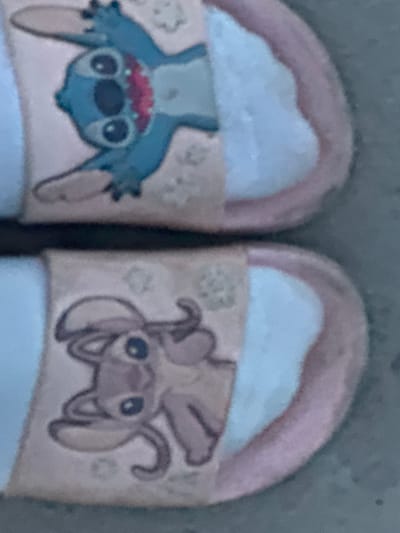 The image shows a pair of pink and white cartoon character slide sandals. The top sandal features a blue cartoon character with large eyes and a red mouth. The bottom sandal shows a light pink cartoon character with large eyes and antennae-like ears. The sandals appear to be on a paved surface, possibly a sidewalk or patio, in an outdoor environment. The location is described as Гожули, Ukraine. The overall impression is of casual footwear with a playful design.