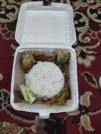 A close-up, overhead shot shows a white styrofoam food container with a meal on a red carpet with a brown and tan patterned design. The food container is open, with the lid lifted upwards and out of frame. Inside the container, a mound of white rice is centrally located, taking up a significant portion of the space. To the left of the rice, a piece of fried chicken is visible. To the right of the rice, a serving of stir-fried vegetables, appearing to be green beans and carrots, is present. In the upper right quadrant of the container, there is a portion of fish with sauce. A few slices of cucumber are placed to the left of the rice. The styrofoam container is resting on a carpet. The image is taken from a high angle, looking down into the food container. No people are visible in the image. The scene is indoors. The food appears to be a take-out meal. The location in Cintadamai, Indonesia is an assumption based on the food presentation and potential cultural context.