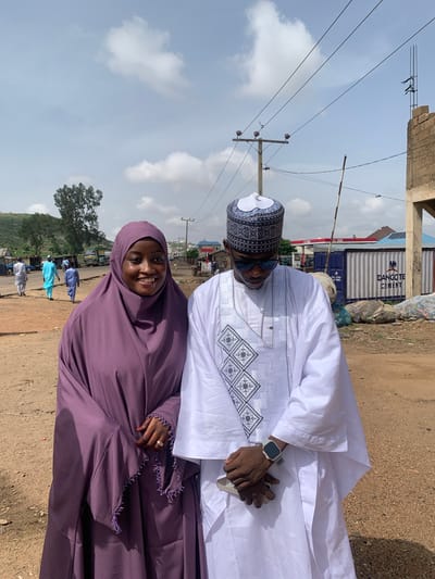 Two individuals are standing close to each other outdoors in Naraguta Hausawa, Nigeria. A woman on the left wears a purple hijab. A man on the right is in white attire, including a patterned hat. He is holding a cell phone and wearing a wristwatch.
In the background, there are utility poles with wires, a building, and shipping containers labeled "DANGOTE CEMENT." Other people are walking in the background. The ground appears to be dirt.