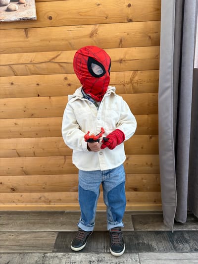 A child dressed in a Spider-Man mask and costume stands on a wooden floor. He wears a red and black Spider-Man mask, a white jacket, blue jeans, and black sneakers with brown laces. He is holding a red toy in his hands. The floor is made of light-colored wooden planks. Part of a gray cushion is visible at the top of the frame. To the right of the wooden floor is a gray and black tiled floor. The location is not discernible in the city of Lermontovo, Armenia from the provided image.