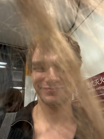 The image shows a close-up, blurry, selfie-style shot of a person with blonde hair and fair skin. The person is smiling and their face is partially obscured by their hair and motion blur. They are wearing a dark-colored, zippered jacket. In the background, there is a window or glass partition, and a sign with red and white text, some of which appears to be Chinese characters and English words like "fire exting". There is also a yellow sticker visible with text. The setting appears to be a public transport vehicle, possibly a subway or train, given the reflective surfaces and signage. The environment is indoors and appears to be well-lit, though the focus is on the individual. The motion blur suggests the image was taken while the subject or the vehicle was in motion. The location is identified as Changning District, China.