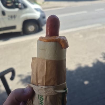 A close-up, waist-up shot shows a person's hand holding a hot dog wrapped in brown paper packaging. The hot dog is topped with an orange sauce and has a dark red casing. In the background, a white van with the letters "3M" on its side is visible, parked on a street with a crosswalk. The scene appears to be outdoors on a sunny day. The image is taken in Bralin, Poland.