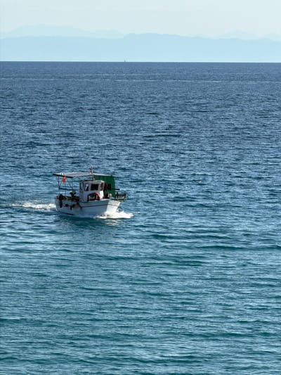 The image depicts a small fishing boat with a white hull and a red stripe, moving through calm blue water. The boat has a cabin, a canopy, and some equipment on deck. A green tarp is visible at the stern. The boat is leaving a white wake behind it, indicating movement. In the background, a hazy mountain range is visible on the horizon, under a pale blue sky. The scene is likely a coastal area of the Municipal Unit of Kastro - Kyllini, Greece, given the clear blue water and mountainous horizon typical of the region. There are no people clearly visible on the boat. The overall impression is one of a serene seascape.