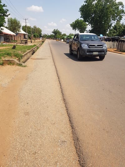 The image depicts a street scene in Palade, Nigeria. A dark-colored SUV is the primary object in the foreground, positioned on the right side of a paved road. The vehicle appears to be stationary. Behind the SUV, the road stretches into the distance, flanked by a drainage channel and vegetation on the left. Further back on the left, there are buildings and trees, suggesting a semi-urban or rural environment. The sky is partly cloudy, indicating daytime. The road surface shows signs of wear and tear. A license plate is visible on the rear of the SUV, bearing the text "FB6-R4". The overall scene suggests a moment captured during a typical day in this Nigerian location.