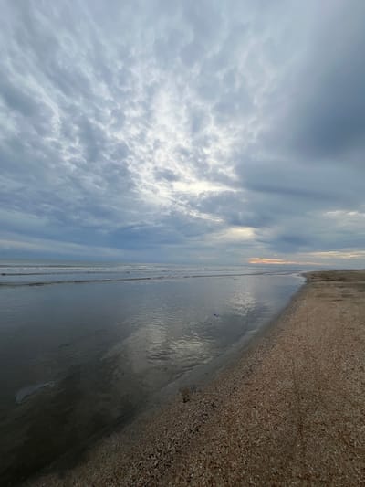 The image displays a coastal landscape in Alueduamuka O, Indonesia, characterized by an overcast sky and a calm sea. The upper two-thirds of the frame are dominated by a dense formation of grey and white clouds, with a concentrated area of bright, diffused light emanating from the upper-left central portion. The horizon line separates the sky from the ocean and shoreline. The sea, occupying the middle-left of the frame, exhibits a dark, reflective surface with gentle, low waves breaking in the distance. The water closest to the viewer reflects the brighter sky, creating a lighter streak. A translucent, elongated object, likely plastic, is partially submerged in the shallow water in the lower-left foreground, and a small, dark blue object floats further out to the right within the water. The beach occupies the lower-right third of the frame, composed of dark, wet sand adjacent to the waterline, transitioning to a wide expanse of drier, light brown sand heavily mixed with small, pale shell fragments. No human individuals, animals, or artificial structures are visible within the scene.