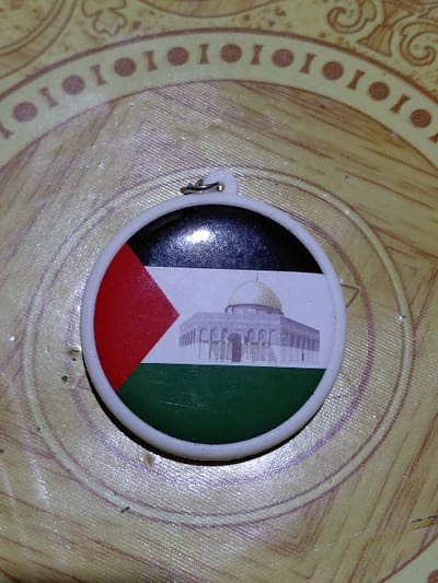 A round, plastic button with the colors of the Palestinian flag—black, white, green, and red—is displayed on a brown surface. The flag is vertically oriented, with the black stripe on the left, followed by white bearing an image of the Dome of the Rock, then green. A red triangle extends from the base. A small, metal loop is visible on the side of the button. The surface beneath has a light brown hue and a circular pattern with dark brown accents.