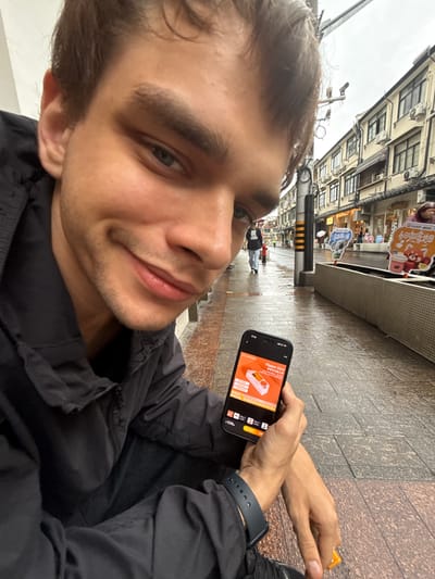 A young man, looking directly at the camera, holds a smartphone displaying an advertisement for "Flipper Zero". He is seated outdoors on a wet sidewalk, with a street lined with traditional Chinese buildings and storefronts in the background. The weather appears overcast, and the ground is damp, suggesting recent rain. A person walks away from the camera in the mid-ground, and other pedestrians and shoppers are visible further down the street. The architecture of the buildings features multi-story structures with awnings and numerous windows, indicative of a commercial area. Signs in Chinese characters are displayed on the storefronts and on street decorations. The overall impression is a snapshot of urban life in Xuhui District, China, on a rainy day.