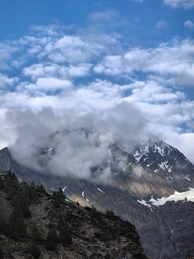 This is a vertical, high-angle shot of a mountainous landscape near Kead, India. The sky is a bright blue, dotted with fluffy white clouds. A tall, rugged mountain dominates the scene, its peak partially obscured by swirling white clouds. Patches of snow are visible on the mountain's upper slopes. In the foreground, a steep, rocky slope is covered with dark green, coniferous trees. Prayer flags, a common sight in this region, are strung along the mountainside. The overall impression is one of natural beauty and spiritual tranquility.