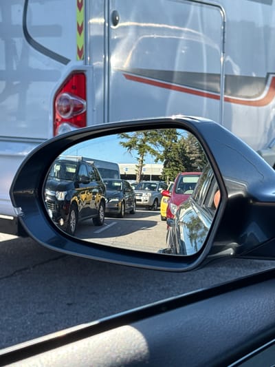 The image shows a rear-view mirror reflecting a line of cars. The cars are stopped, suggesting traffic is at a standstill. In the foreground, the side of a dark-colored vehicle is visible, as is part of the dashboard. The reflection in the mirror reveals a black SUV, a dark sedan, a red car, and a yellow car, all appearing to be in a queue. Beyond the cars, trees and buildings are visible under a clear blue sky. The overall scene suggests a typical traffic situation. The metadata indicates the image was taken in Kapitan Andreevo, Bulgaria.