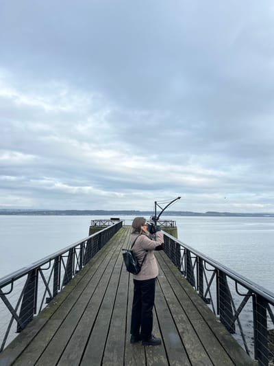 An adult female stands on a wooden pier in Blackness, United Kingdom, under an overcast grey sky. The individual, wearing a light brown textured jacket, dark trousers, and dark boots, with a black backpack on her left shoulder, is positioned centrally on the pier, facing away from the viewer. She holds a dark imaging device, possibly binoculars or a camera, to her eyes, looking out over the calm, grey water. Dark metal railings with vertical and diagonal supports line both sides of the pier. Towards the distant end of the pier, a dark, skeletal metal structure resembling a crane is visible. Across the water, a distant landmass with some discernible features stretches horizontally. A small, dark bird is captured in flight over the water to the right, in the mid-distance.