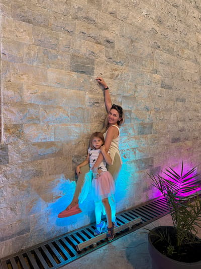 A woman and a young girl are posing against a textured stone wall illuminated by blue and pink lights. The woman, with blonde hair and sunglasses perched on her head, is wearing a cream-colored tank top and tan pants. She is leaning against the wall, with her right arm extended upwards. The girl, in a striped top and a pink tutu, stands in front of the woman, looking towards the camera. Her left arm is around the woman's waist. Both are wearing shoes, with the woman in pink platform sandals and the girl in blue sandals. A metal grate runs along the base of the wall, with a potted palm plant to the right, casting a vibrant purple glow. The scene appears to be an outdoor or semi-outdoor space in the city of Zagora, Montenegro, possibly a courtyard or patio area.