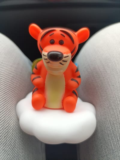 An orange and black plastic figurine of Tigger, from Winnie the Pooh, sits on a white cloud-shaped object. The background consists of two gray objects that resemble clothing, and a blurry black space. There is no indication of this image being in Kirsanovka, Russia.