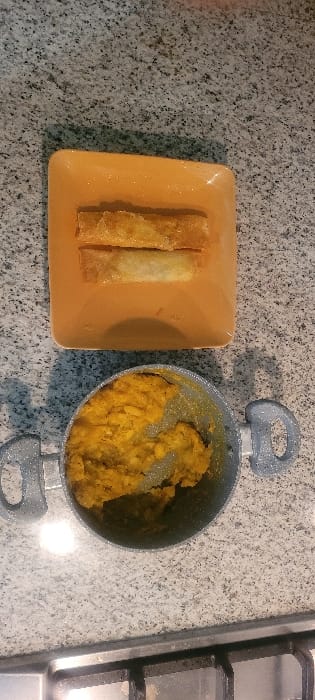 Three spring rolls rest on a square orange plate. A grey pot filled with yellow filling sits beside the plate. The pot has two handles, and the filling appears mashed. A portion of a stovetop burner is visible to the right of the pot. All of these items are placed on a speckled grey countertop.