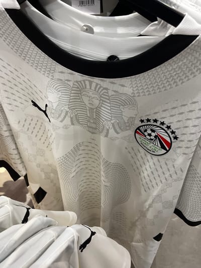 The image is a close-up, eye-level shot of a white football jersey with a black collar. The jersey features a subtle, repeating pattern in a lighter shade of gray, incorporating geometric shapes and what appear to be stylized Egyptian pharaoh heads. On the left side of the chest, a black Puma logo is visible. On the right side of the chest, an oval-shaped emblem is present, containing a soccer ball, stars, and Arabic text which translates to "Egyptian FA". The jersey is hanging, and part of another white jersey is visible in the foreground, also bearing a black Puma logo on its sleeve. The background is out of focus but suggests a retail environment, possibly a sports apparel store, with clothing racks and other garments. There are no people or specific landmarks indicating a location in Rodengo, Italy. The lighting is even, illuminating the details of the jersey.