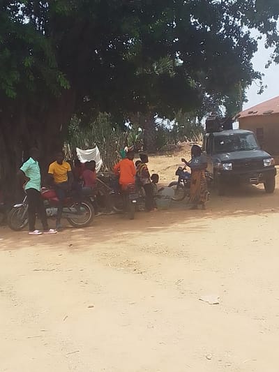 The image depicts a scene in Kwal, Nigeria, at coordinates 9.804 latitude and 8.647 longitude. A group of people, including adults and children, are gathered under a large tree. They are standing around and sitting on several parked motorcycles. One person, wearing a yellow shirt, is sitting on a motorcycle, while another person, wearing a teal shirt, is standing next to it. There is a large black truck parked to the right of the group of people. The truck is facing away from the camera and has a white cloth draped over its rear window. The people are interacting with each other and appear to be having a conversation. The environment is dusty and dry with sparse vegetation. The background includes a building with a red roof and several trees. The scene appears to be taking place on a dirt road.