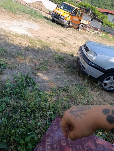 The image depicts an outdoor scene in Gorna Breznitsa, Bulgaria, likely during a sunny day. The foreground features a hand with tattoos, resting on a piece of wooden board with "GTC FORMWORK" and "TARTA INDONESIA" printed on it. In the mid-ground, a silver Volkswagen car is parked. Behind the car, an orange and yellow Iveco truck is visible, its door open, and with "cmpouzpyn" and a phone number printed on it. The truck appears to be involved in some form of construction or material transport, with wooden planks and a pile of dirt visible behind it. The setting is somewhat rural, with dry grass and scattered greenery, and hints of houses with red roofs and trees in the background. The overall impression is of a construction site or a place where such vehicles and materials are stored. The sunlight suggests it is daytime, likely late morning or afternoon.