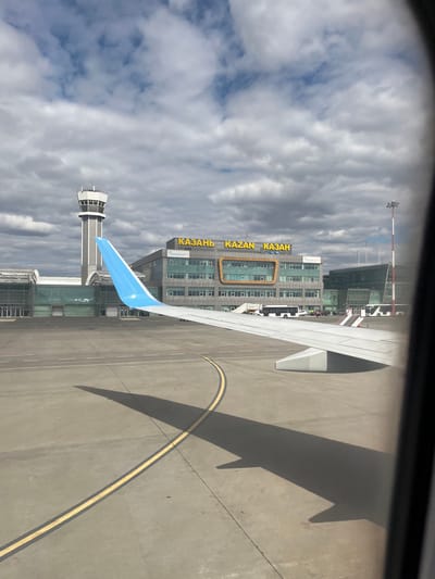 The image shows a view from an airplane window, likely at Kazan International Airport in Песчаные Ковали, Russia. An airplane wing is visible, with the shadow cast on the tarmac. The airport control tower and the main airport building with the word "KAZAN" in large gold letters are visible. The sky is overcast with clouds.