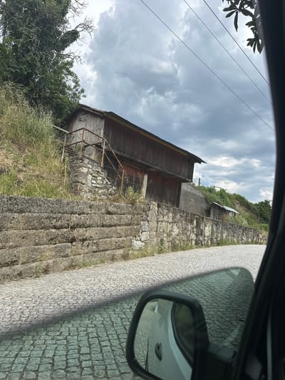The image shows a view from inside a vehicle, capturing a street scene in Amarante, Portugal. A weathered wooden building with a rusty metal railing sits adjacent to a stone retaining wall with integrated steps. The stone wall runs alongside a narrow cobblestone street. Overcast skies are visible in the background. A side mirror of the vehicle is in the frame, reflecting the street. Vegetation grows around the base of the building and along the wall.