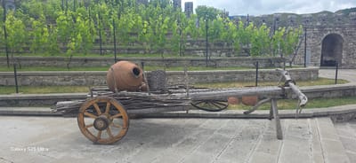 An outdoor scene depicts a historical setting with a rustic wooden cart in the foreground, laden with branches and an ancient clay amphora. The cart has one large wooden wheel visible on the left. Behind the cart, tiered stone walls support a vineyard with lush green grapevines. In the background, a stone fortress with arched openings and battlements overlooks the landscape, with rolling hills under a cloudy sky. The cart and amphora are positioned on a paved surface. The "Galaxy S25 Ultra" watermark is visible in the lower left corner of the image. This scene is located within the walls of the Rabati Castle complex in Akhaltsikhe, Georgia.