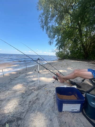The image depicts a serene fishing scene on the sandy shore of the Kyiv Reservoir near Kozarovychi, Ukraine. In the foreground, a person is seated in a camping chair, their legs propped up, wearing blue patterned shorts and barefoot. Two fishing rods are positioned on the sand, supported by a tripod and another rod holder. A blue bucket containing fishing bait sits nearby, along with a dark green fishing bag. The background reveals the calm waters of the reservoir under a clear blue sky, with gentle waves lapping the shore. Lush green trees line the distant bank, casting dappled shadows on the sandy beach. The overall atmosphere is one of peaceful relaxation and outdoor recreation.
