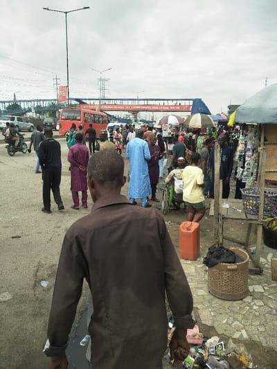 The image captures a bustling outdoor market and roadside scene in Ajah, Nigeria, specifically near the Abraham Salewa and VET, 3rd HFP Complex, Block F3, as indicated by signage on an elevated pedestrian bridge. The sky is overcast. A worn, grey asphalt road occupies the left and central mid-ground, transitioning to a more uneven paved and dirt market area on the right.
Approximately 30-40 individuals are present. In the immediate foreground, an adult male, viewed from behind, wears a plain dark brown long-sleeved shirt and walks towards the market. In the midground, a dense crowd of pedestrians traverses both the road and market area. Notable individuals include a man in a purple traditional garment, a man in a light blue agbada, a person in a yellow t-shirt, and a person in a wheelchair within the market crowd. On the far left, a person on a motorcycle is visible on the road.
Vehicular traffic includes a red public transport bus (BRT type) in the mid-background, multiple cars, and a motorcycle. Overhead, power lines and a tall lamppost are visible. The right side of the image features numerous makeshift market stalls, constructed with various coverings such as blue tarps, striped umbrellas, and grey awnings. Merchandise, including packaged goods and bananas, is displayed. An orange plastic jerrycan and a woven basket are on the ground near the stalls. Significant litter, consisting of plastic wrappers and other debris, is accumulated on the ground in the foreground right.