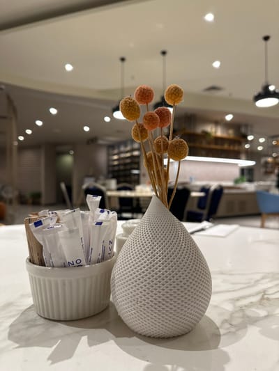 The scene depicts a tabletop in what appears to be a café or restaurant located in Zeytinburnu, Turkey. In the foreground, a white marble-patterned table holds two primary objects. To the left, a white, ribbed ceramic ramekin contains numerous individually packaged sugar or sweetener packets, some white with blue text (partially visible "UNO" and "NTN"), others brown. To the right, a teardrop-shaped decorative vase, light grey with a prominent grid-like texture, holds a cluster of dried or artificial stems topped with small, round, fuzzy spheres in shades of orange and yellow. Shadows are cast on the table surface from both objects. The background, out of focus, reveals an indoor commercial space with recessed ceiling lights and several black pendant lights. Tables with dark blue and lighter chairs are visible, along with a long counter or serving area and shelving units. No individuals are present in the image.