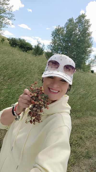 A woman in a yellow hoodie and a white, jeweled baseball cap, wearing sunglasses, is holding a bunch of wild strawberries in her left hand. She is smiling at the camera. The background is a grassy hillside with trees under a cloudy blue sky. The location is the city of Pervomayskoye, Russia.
