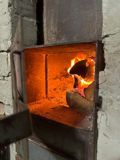 The image shows the interior of a rustic fireplace in the city of Сароцкі сельскі Савет, Belarus. A fire is burning brightly, with flames consuming logs of wood. The fireplace is made of metal and stone, and the surrounding walls are made of rough, textured material. The open door of the fireplace is visible on the left side of the image. Ash and embers are scattered on the floor of the fireplace.