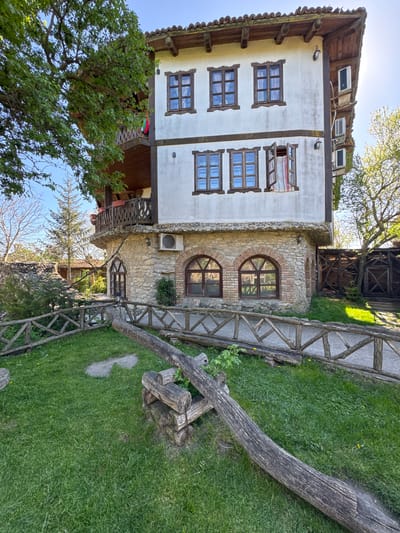 The image shows a building in Kalimantsi, Bulgaria. It features a two-story structure with a white facade, brown trim around the windows, and a red-tiled roof. The building is constructed with a combination of white plaster and stone. There are multiple windows visible on the facade, some of which have open shutters. The building is also equipped with air conditioning units.

The building is surrounded by greenery, including grass, trees, and shrubs. There's a wooden fence along a pathway. A large tree trunk rests in the foreground. A wooden structure is situated between the main building and the fence.