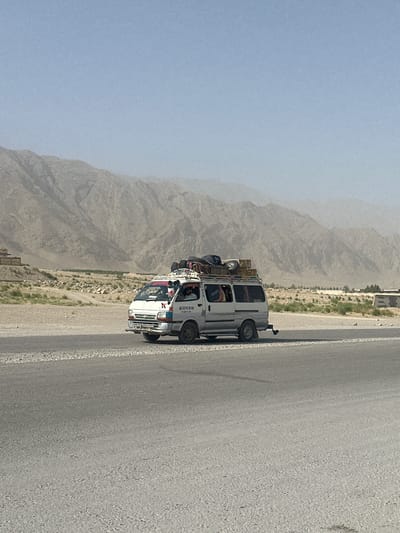 A white van, heavily laden with luggage and cargo on its roof, travels along a paved road. The van has several people inside and appears to be in motion. The surrounding environment is arid, with dry vegetation and dusty mountains in the background. The sky is a hazy blue. The van's markings suggest it might be a form of public transport or commercial vehicle. The location is identified as the city of Bostan, Pakistan.