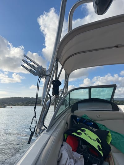 The image depicts a forensic scene aboard a motorboat traversing a body of water, likely the Minho River, within or near Vila Nova de Cerveira, Portugal. The perspective is from within the boat, looking forward and to the port side.
In the foreground, a padded, white upholstered boat seat is visible, marked with the number "215" on its side. On this seat, various items are stored: a black and neon yellow personal flotation device or impact vest, a red baseball cap, white folded textiles (possibly towels or clothing), and a black wakeboarding glove.
Dominating the left side of the frame is a polished metal wakeboard tower supporting a white canvas bimini top, which provides shade. Attached to the tower is a silver-colored, three-pronged wakeboard rack, designed to hold a wakeboard. A black glove hangs from the tower, and black cables or ropes are visible, dangling from the tower structure. A black, cylindrical object, likely a speaker or light, is also mounted on the tower.
To the right, a tinted boat windshield with a walk-through opening is visible, suggesting the boat's forward cabin or bow area.
Beyond the boat's immediate structure, a body of water shows ripples and a distinct wake trailing behind the vessel, indicating it is in motion. In the mid-ground on the water, a red buoy is present, marking a channel or hazard. The background features a distant, undulating green shoreline densely covered with vegetation and hills, under a blue sky with scattered white cumulus clouds. No individuals are visible in the scene.