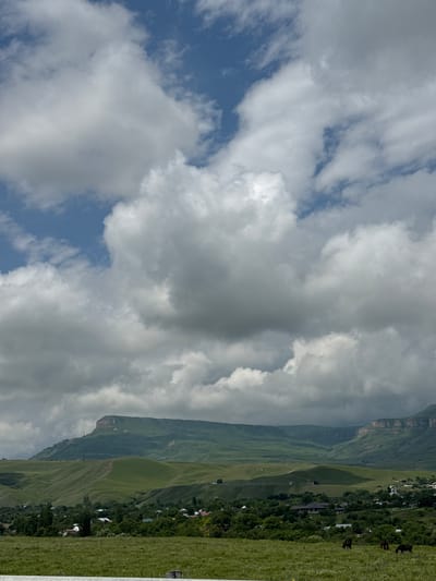 A wide, landscape view captures a rural scene in the village of Kubina, Russia, under a cloudy sky. The foreground features a grassy field with two horses grazing. In the midground, a village is nestled amongst trees and rolling green hills. The background is dominated by a large, flat-topped mountain range, its slopes also covered in green vegetation. The sky is a mix of blue patches and large, dramatic white and gray clouds.