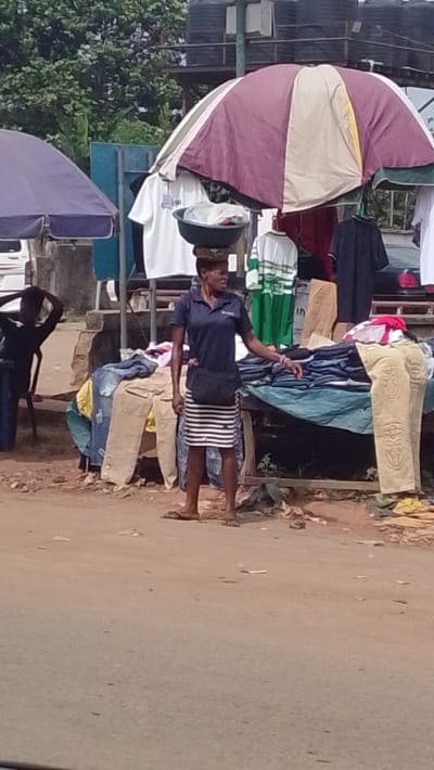 The scene depicts an outdoor street market or vending area in Igbo Ukwu, Nigeria. In the foreground, an adult woman stands slightly right of center, facing right. She wears a dark blue polo shirt, a black and white horizontal striped skirt, and light-colored flip-flops, with a black fanny pack strapped across her torso. A black basin containing items, including a white plastic bag, is balanced on her head. Her left hand rests on a pile of folded clothing displayed on a wooden table covered by a greenish-blue tarpaulin. Behind her and to her right, a large market umbrella with alternating dark red, beige, and green sections provides shade over additional clothing displays. Various shirts are hanging from the umbrella's structure and a stand behind it, including white t-shirts and a green and white striped shirt. To the woman's right, more folded clothes, including a pair of light brown pants with an embossed pattern, are visible on another draped table. On the far left, partially obscured by a blue market umbrella, another individual sits on a dark chair with their hands behind their head. The ground is unpaved dirt. In the background, green foliage is visible, along with concrete structures and multiple dark water tanks elevated on a platform. A portion of a white vehicle is also visible on the far left.