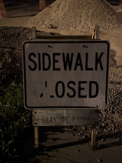A nighttime, eye-level, close-up shot shows a weathered metal sign that reads "SIDEWALK CLOSED." The sign is supported by two wooden posts attached to a metal frame. Below the primary sign, another metal plate is visible with embossed text that appears to read "CITY OF PARMA." The background consists of a pile of grey gravel and dark soil, with some green grass and foliage visible on the left side of the frame. Orange construction cones and what appear to be piles of dark asphalt are faintly visible in the upper portion of the image, suggesting ongoing construction work. The overall scene is dark, with artificial lighting casting shadows.