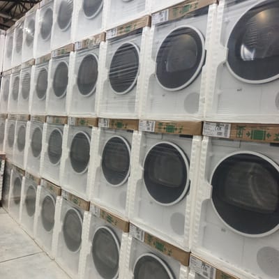 The image shows a large stack of new, white Bosch clothes dryers packaged in plastic wrap and foam. The dryers are arranged in multiple rows and columns, creating a warehouse-like setting. Some of the packages have brown cardboard bands with visible text, including "BOSCH," "031 V08," "CEERC," "AN88211ME," and "max. 1000kg." The setting appears to be indoors, likely a retail or storage facility, with a glimpse of a metal ceiling structure at the top of the image. The lighting is even, suggesting artificial illumination, and there are no people visible in the frame. The scene focuses solely on the stacked appliances, highlighting their uniformity and packaging. The location is identified as Britovo, Russia.