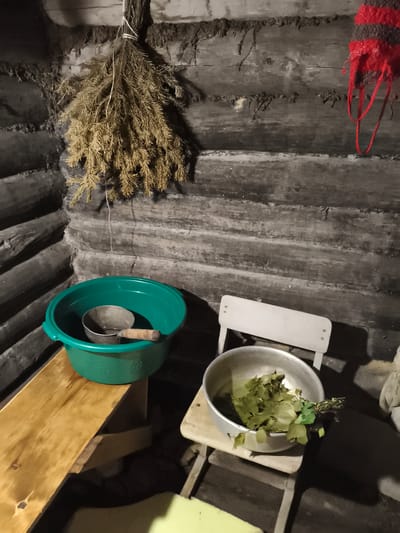 The image depicts an indoor corner of a rustic wooden structure, likely a sauna or bathhouse, in Il'mino, Russia. The walls are constructed from dark, weathered logs, horizontally stacked. Hanging from the upper left wall is a bundle of dried fir branches, tied with white string. To the right, a portion of a red and black striped fabric, possibly a rug or decoration, is visible.

In the foreground, a green plastic basin sits on a wooden bench. Inside the basin is a smaller metal basin containing water and a metal ladle with a wooden handle. To the right of the green basin, a simple, light-colored folding chair is positioned. On the seat of the chair rests a large metal bowl filled with fresh green leaves, resembling a bath whisk or venik. The floor appears to be made of dark wood. The lighting is dim, creating shadows and highlighting the textures of the wood and objects. No individuals are present in the scene.
